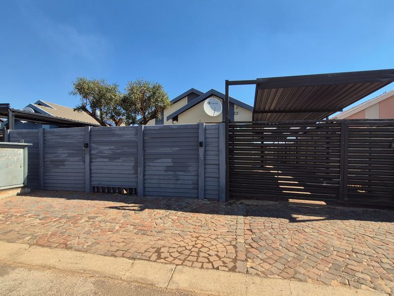 Front exterior view showing a paved driveway, secure slatted metal gate, and a covered carport structure.
