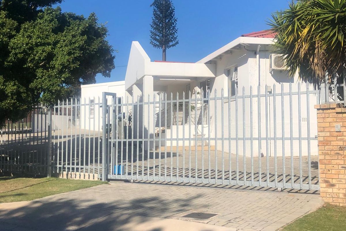 Front exterior view of a commercial property featuring a secure steel gate, paved driveway, and white painted facade with an installed air conditioning unit.