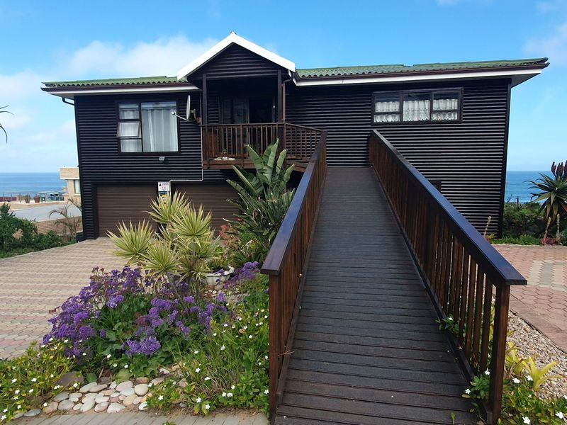Front elevation of a coastal home featuring dark corrugated metal cladding, a prominent timber access ramp, and a paved driveway with ocean views.