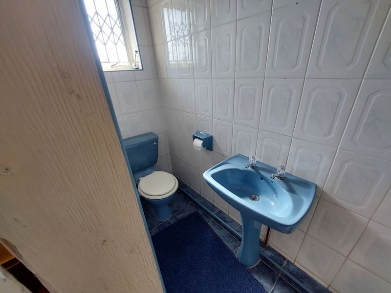A compact bathroom featuring dated blue sanitaryware, including a pedestal basin and toilet, with full-height wall tiling and a security window gate.