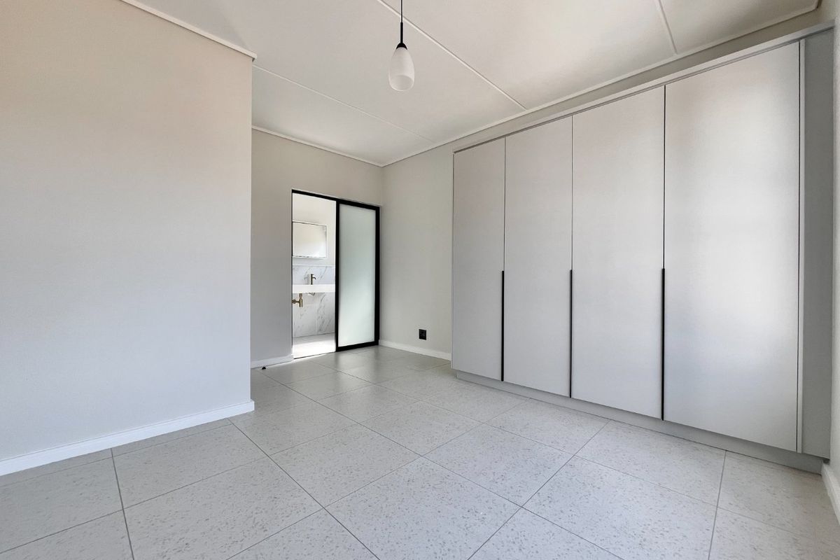 A bright, modern bedroom featuring neutral tiled flooring, ample built-in cupboard space, and direct access to an en-suite bathroom.