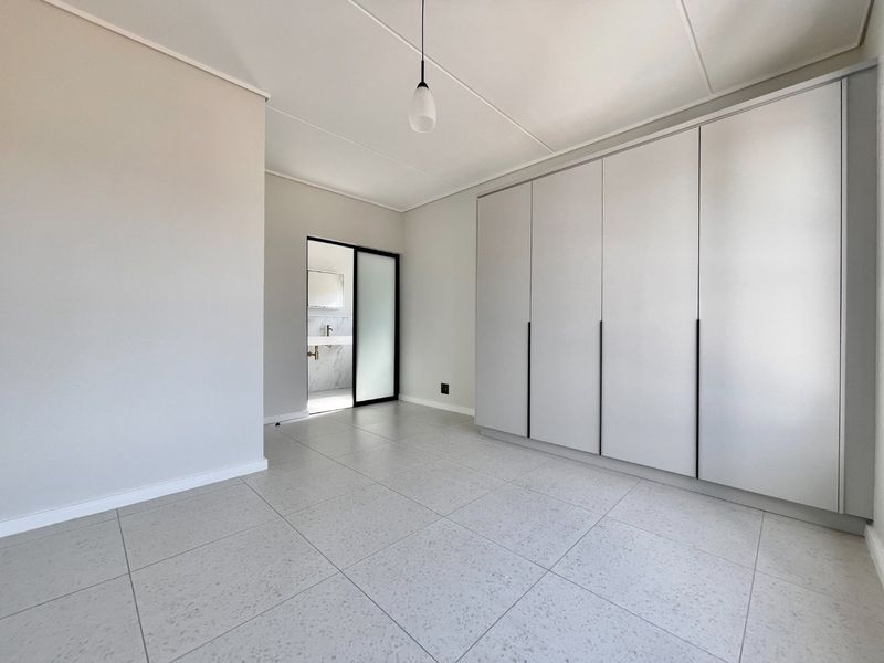 A bright, modern bedroom featuring neutral tiled flooring, ample built-in cupboard space, and direct access to an en-suite bathroom.