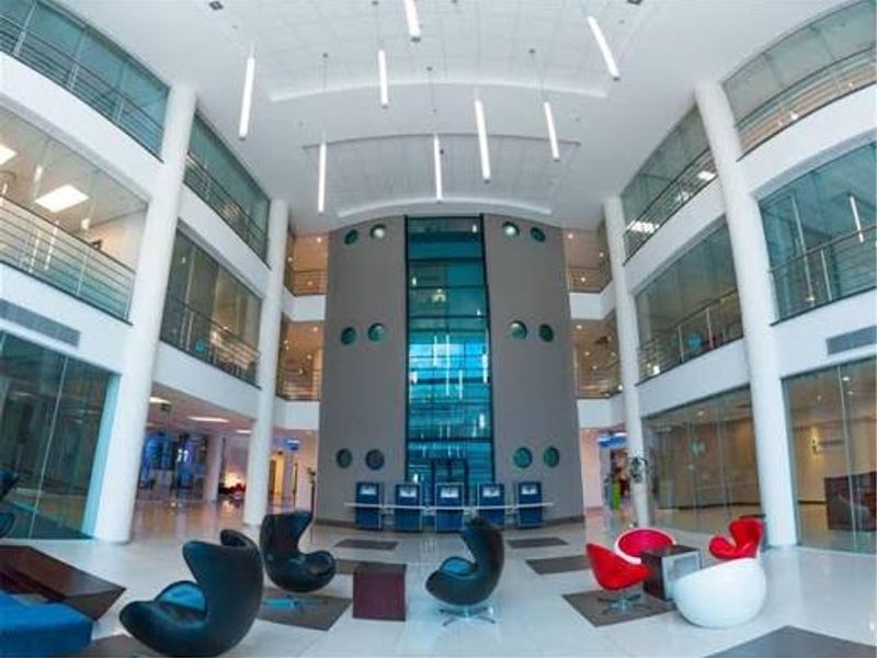 Spacious modern commercial atrium featuring high ceilings, glass-enclosed office partitions, a central glass elevator shaft, and polished tile flooring with contemporary lighting.