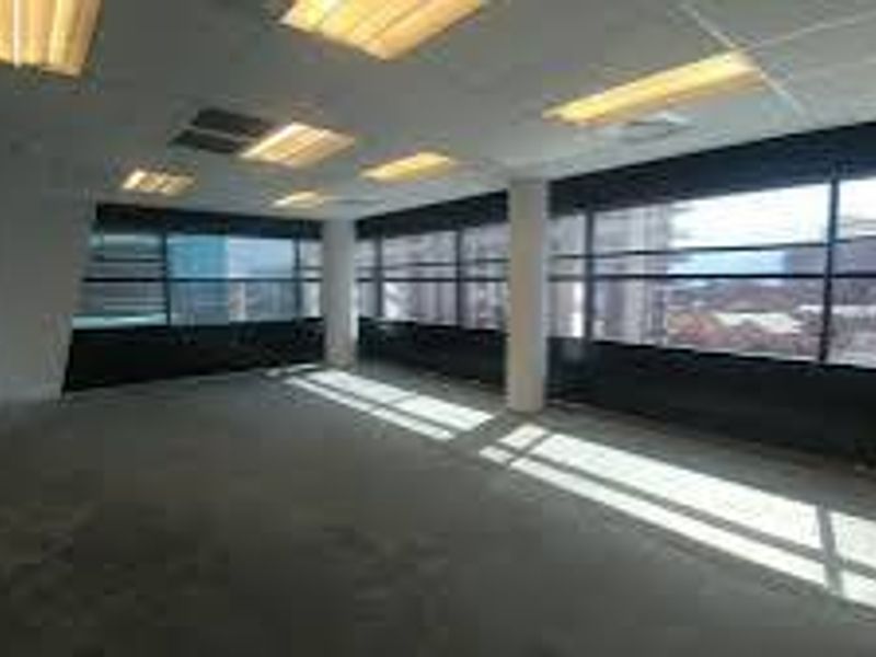 An empty, open-plan commercial office space featuring large windows, structural columns, and a suspended ceiling with integrated lighting.