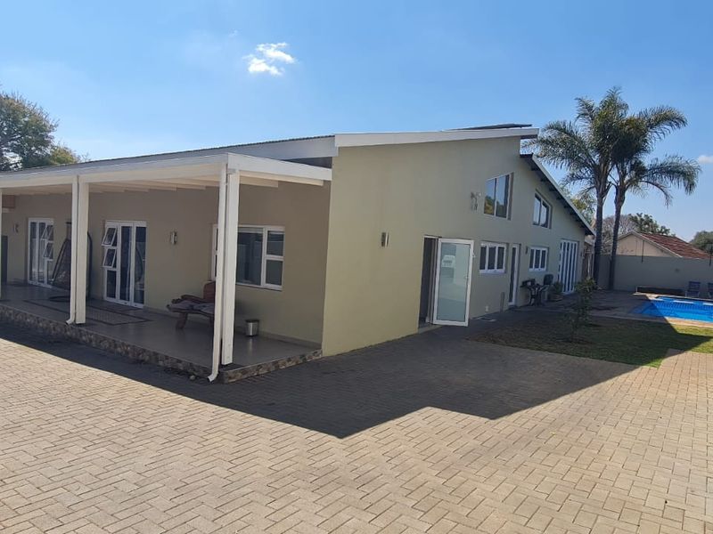 Rear view of the property featuring a spacious paved courtyard, a covered patio area, and a swimming pool set within a landscaped garden.