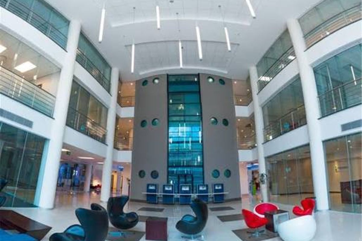 Spacious modern commercial atrium featuring high ceilings, glass-enclosed office partitions, a central glass elevator shaft, and polished tile flooring with contemporary lighting.