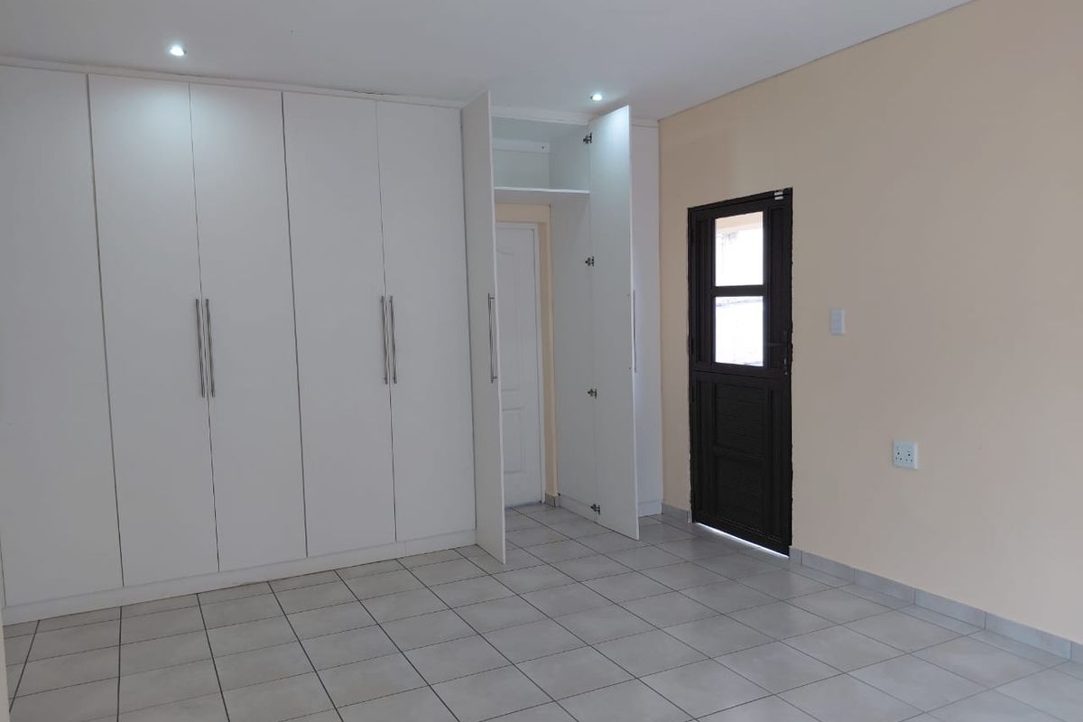 A clean, empty bedroom featuring neutral tiled flooring, extensive white built-in cupboards, recessed lighting, and direct access to an exterior door.
