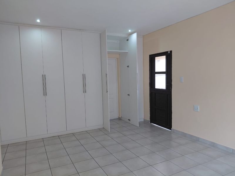 A clean, empty bedroom featuring neutral tiled flooring, extensive white built-in cupboards, recessed lighting, and direct access to an exterior door.