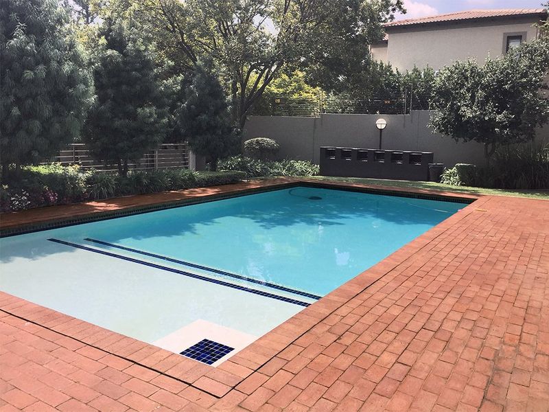 A well-maintained swimming pool area featuring brick paving, established garden landscaping, perimeter wall with electric fencing, and a built-in outdoor braai facility.