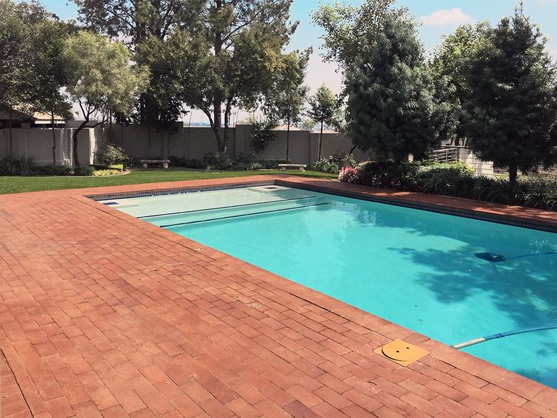 A well-maintained swimming pool area featuring brick paving, a pool cover, and a landscaped garden with a perimeter boundary wall.