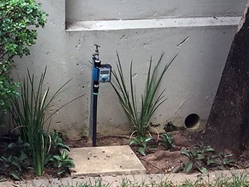 A close-up view of an external water tap fitted with an irrigation timer, located against a concrete wall in a garden setting.