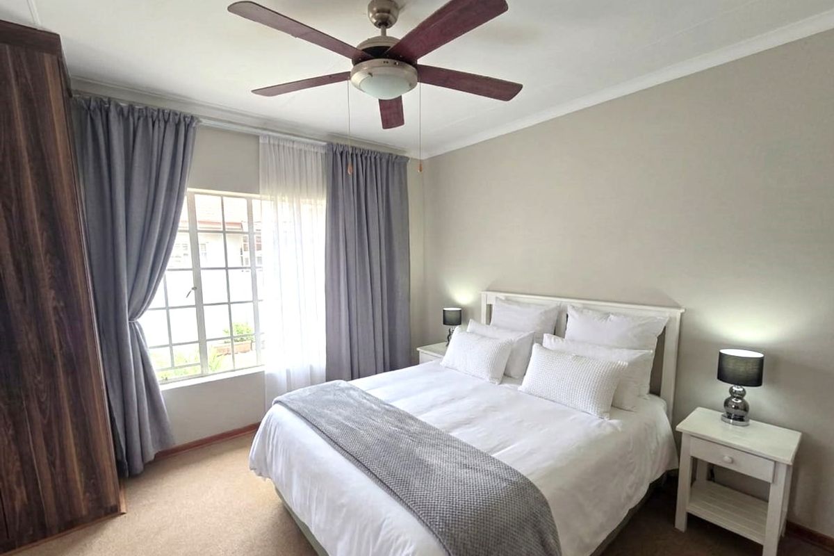 A well-maintained bedroom featuring a ceiling fan, built-in wardrobe, and large window with security bars, finished with neutral carpeting.