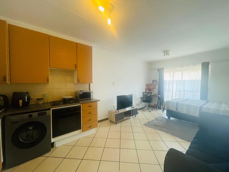 Open-plan studio apartment featuring a compact kitchen with built-in cabinetry, oven, and hob, tiled flooring, and access to a balcony through sliding glass doors.