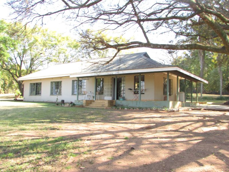 A single-story farmhouse featuring a covered veranda, metal roofing, and steel-framed windows, set within a spacious, tree-lined garden area.