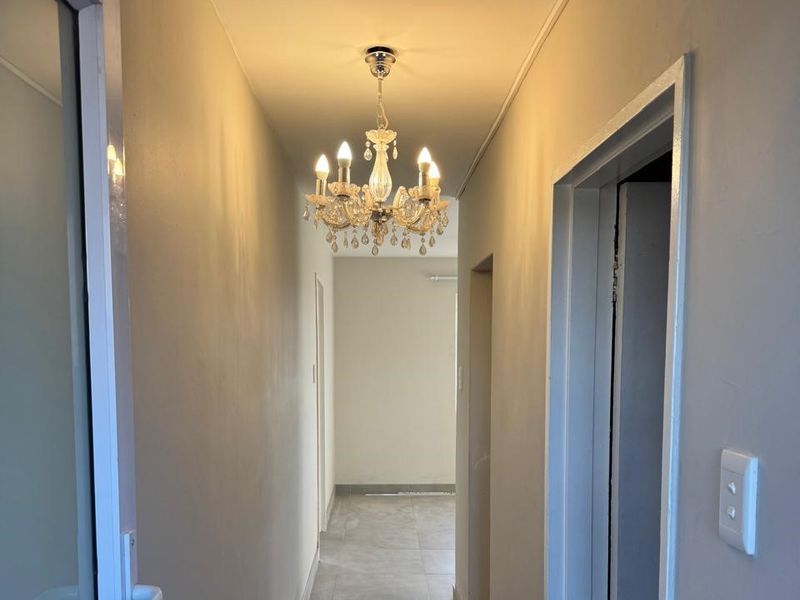 A clean, well-lit hallway featuring neutral tiled flooring and a decorative chandelier, providing access to adjacent rooms within the apartment.