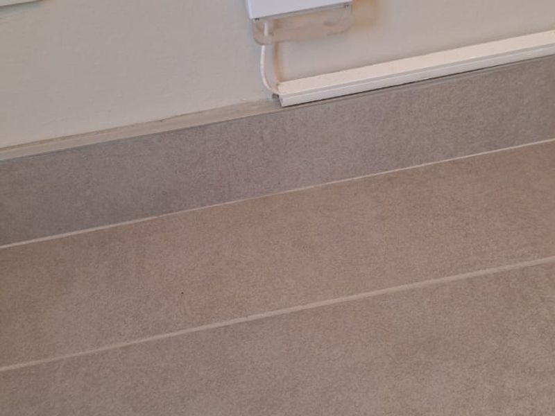 Close-up view of tiled flooring, skirting board, and a wall-mounted electrical component in an apartment.