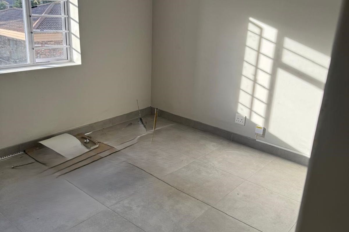 An empty bedroom featuring large format floor tiles, a decorative chandelier, and security bars on the window, currently requiring minor repairs to flooring and wiring.