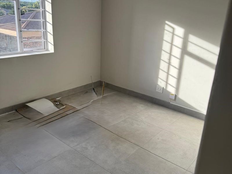 An empty bedroom featuring large format floor tiles, a decorative chandelier, and security bars on the window, currently requiring minor repairs to flooring and wiring.