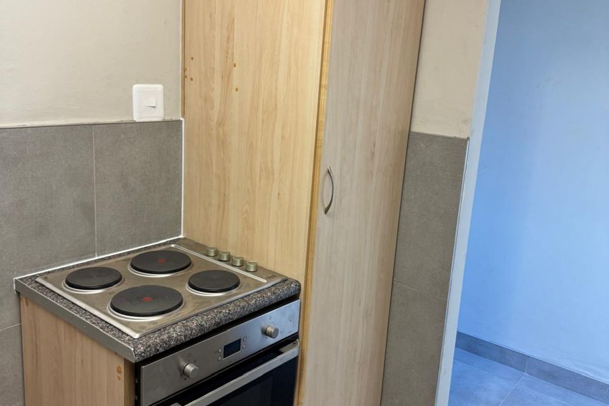 A compact kitchen area featuring a built-in oven, electric hob, and a tall pantry cupboard with grey tiled splashbacks and flooring.
