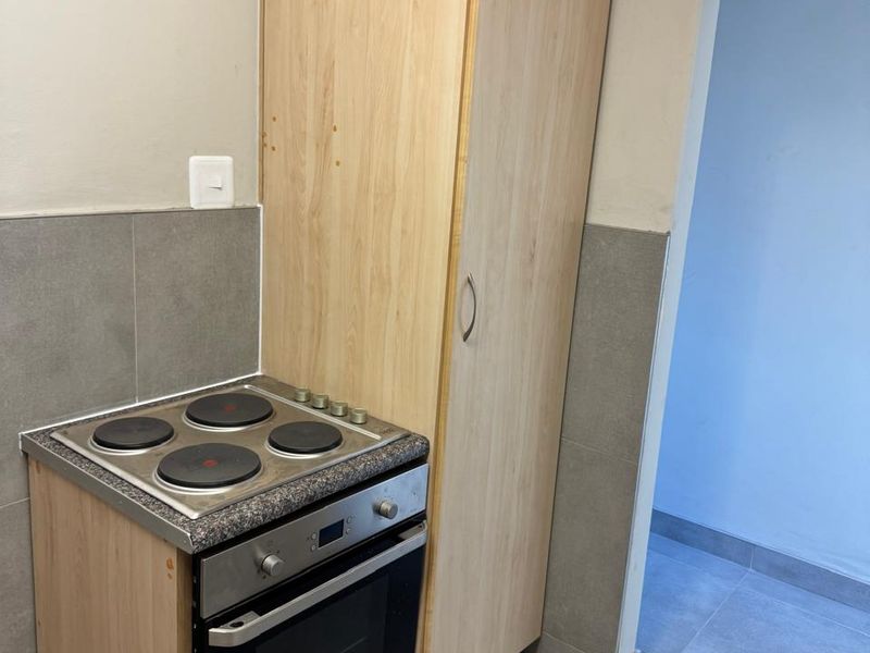 A compact kitchen area featuring a built-in oven, electric hob, and a tall pantry cupboard with grey tiled splashbacks and flooring.