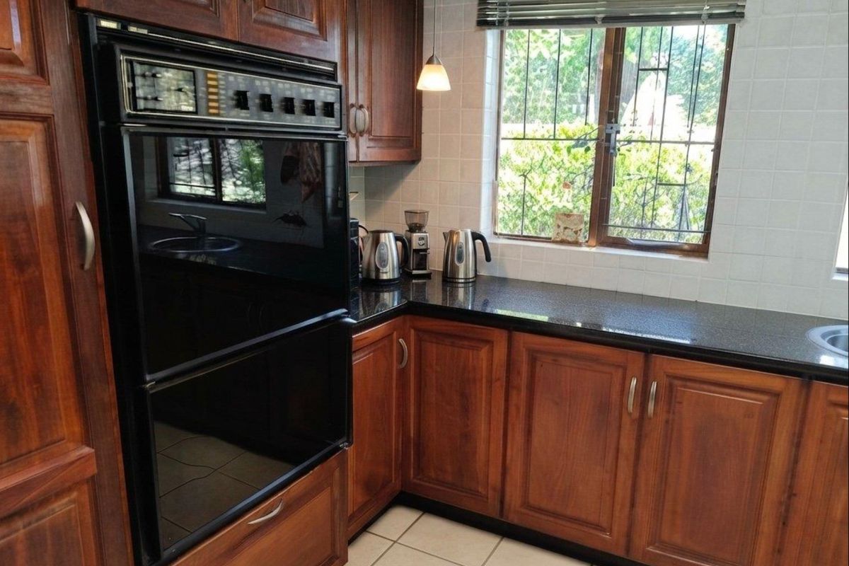 A functional kitchen featuring dark wood cabinetry, granite countertops, a built-in double oven, and a window with security bars overlooking the garden.