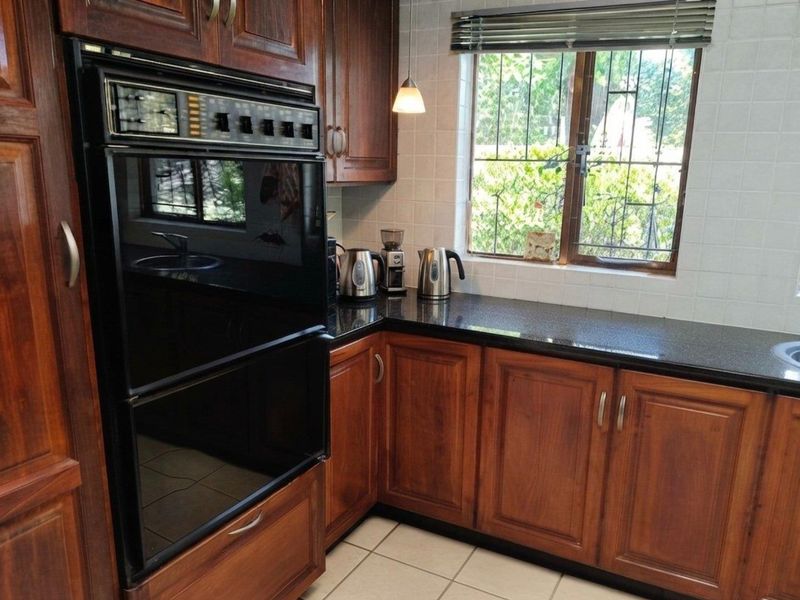 A functional kitchen featuring dark wood cabinetry, granite countertops, a built-in double oven, and a window with security bars overlooking the garden.