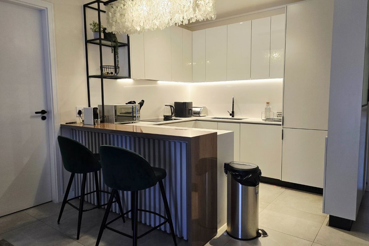 Modern kitchen featuring sleek white cabinetry, stone countertops, under-cabinet lighting, and a functional breakfast bar with integrated shelving.