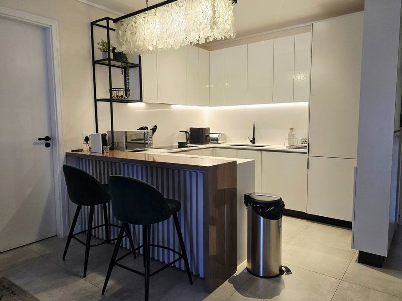 Modern kitchen featuring sleek white cabinetry, stone countertops, under-cabinet lighting, and a functional breakfast bar with integrated shelving.