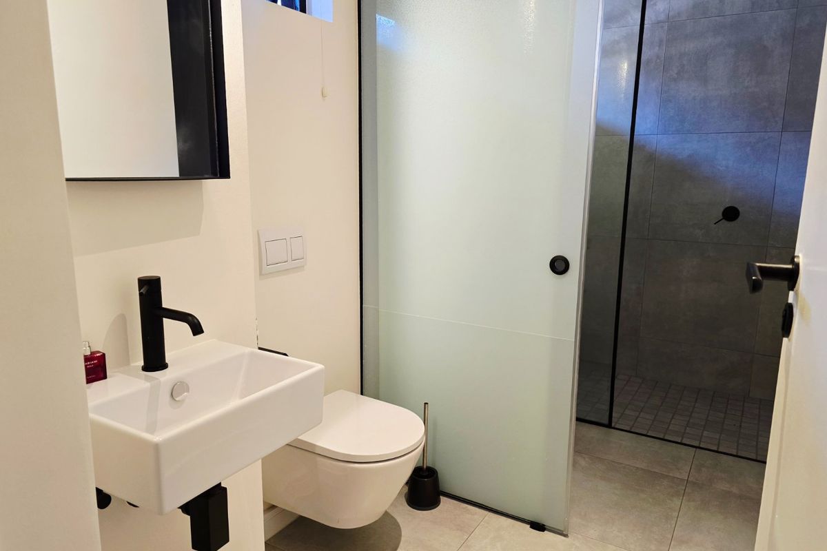 Modern bathroom featuring a wall-mounted basin with black fixtures, a wall-hung toilet, and a spacious walk-in shower with a frosted glass door.