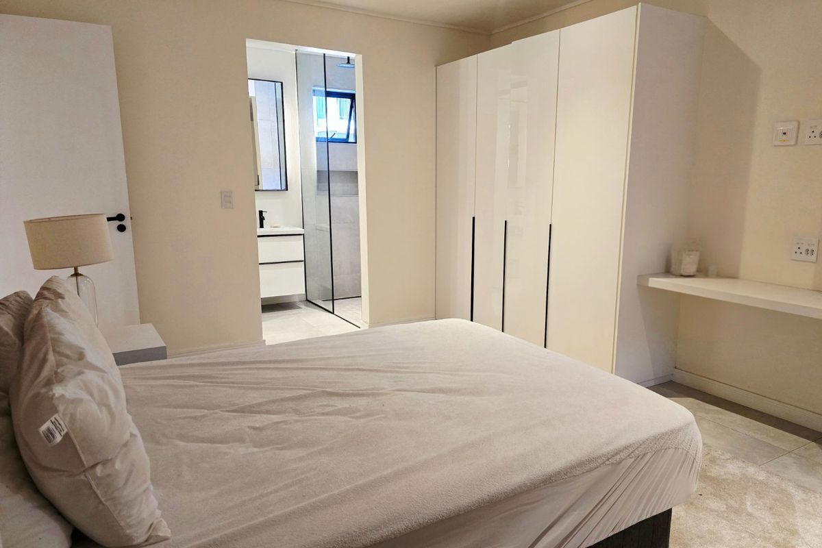 A modern bedroom featuring a built-in wardrobe, a floating desk shelf, and an adjoining en-suite bathroom with a glass-enclosed shower.