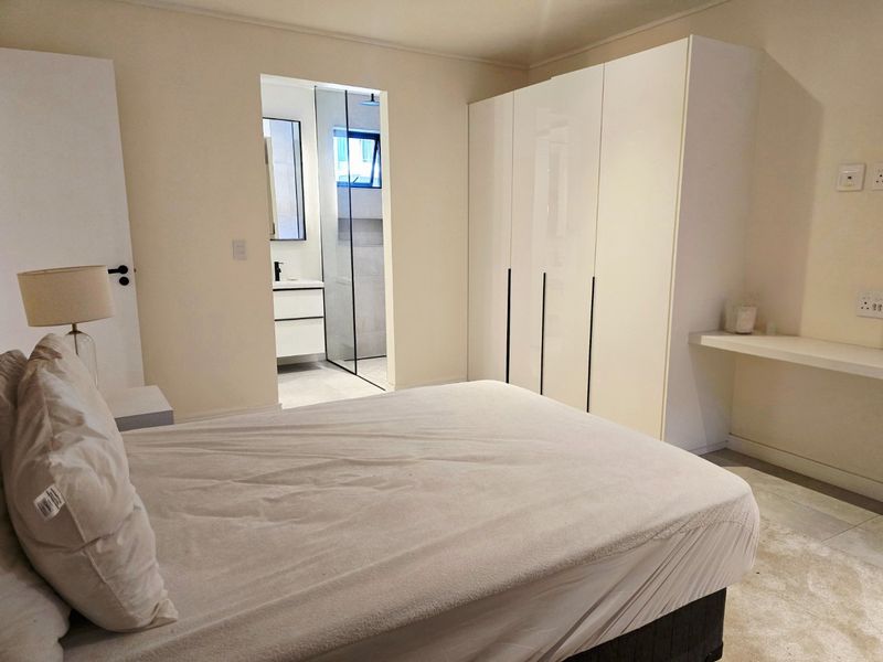 A modern bedroom featuring a built-in wardrobe, a floating desk shelf, and an adjoining en-suite bathroom with a glass-enclosed shower.