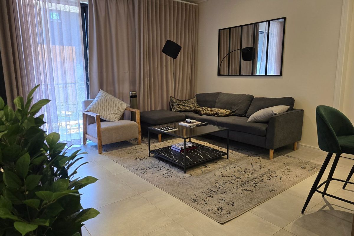 A well-maintained living room featuring large sliding glass doors with floor-to-ceiling curtains, neutral tiled flooring, and a decorative wall mirror.