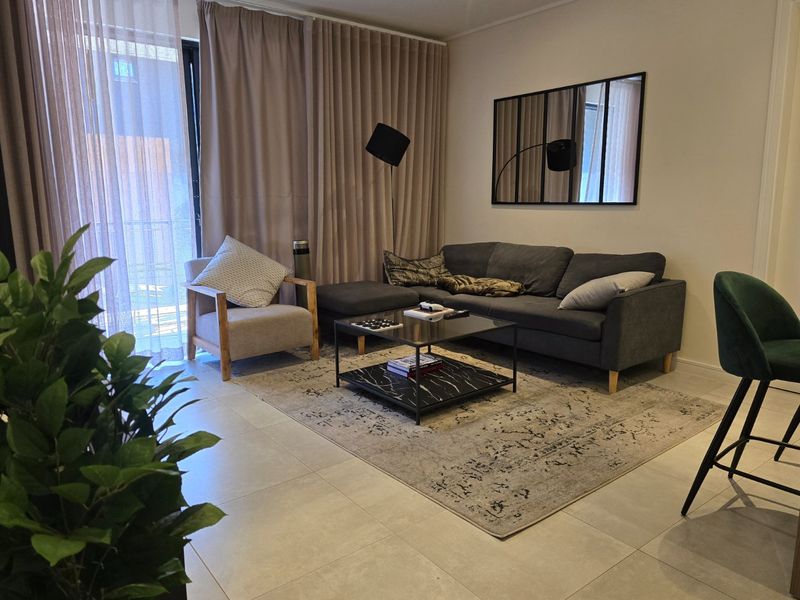 A well-maintained living room featuring large sliding glass doors with floor-to-ceiling curtains, neutral tiled flooring, and a decorative wall mirror.