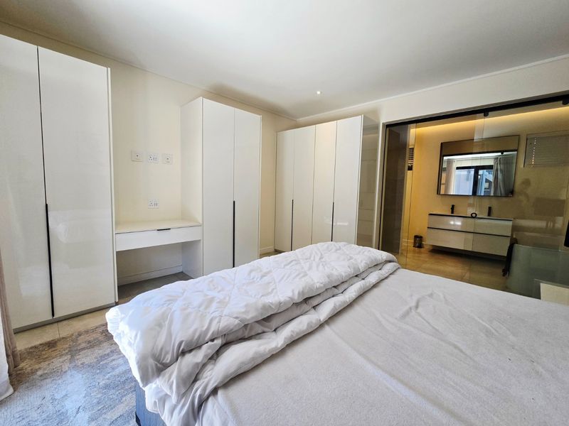 Modern bedroom featuring extensive built-in white cabinetry, integrated vanity, and a sleek glass partition wall leading to an en-suite bathroom.