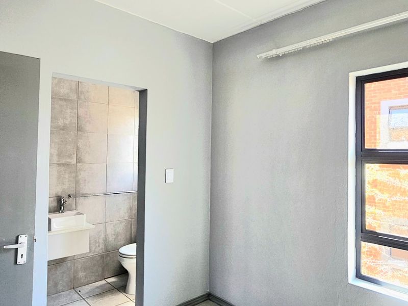 A clean, empty bedroom featuring neutral tiled flooring, grey walls, and an adjoining en-suite bathroom with modern tiling.