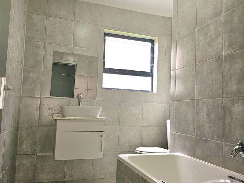 A modern, fully tiled bathroom featuring a wall-mounted vanity with a vessel sink, a bathtub, and a toilet, finished with neutral grey tiles.