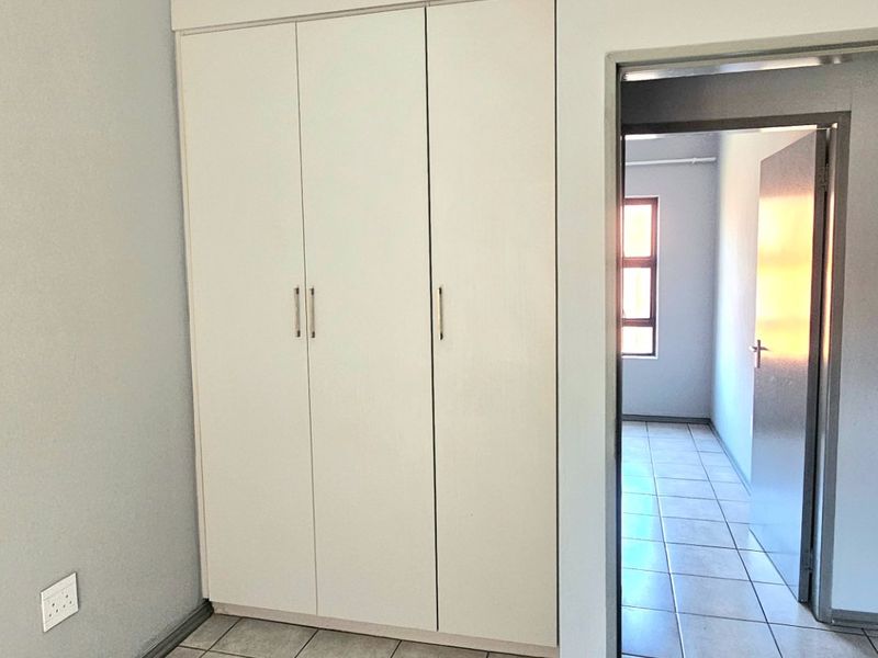 A clean, empty bedroom featuring neutral grey walls, tiled flooring, and built-in white cupboards, with a view into the adjacent hallway.