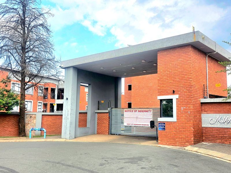 Front entrance view of Mikkadesh apartment complex featuring a secure gated access point with brick facade and electric fencing.