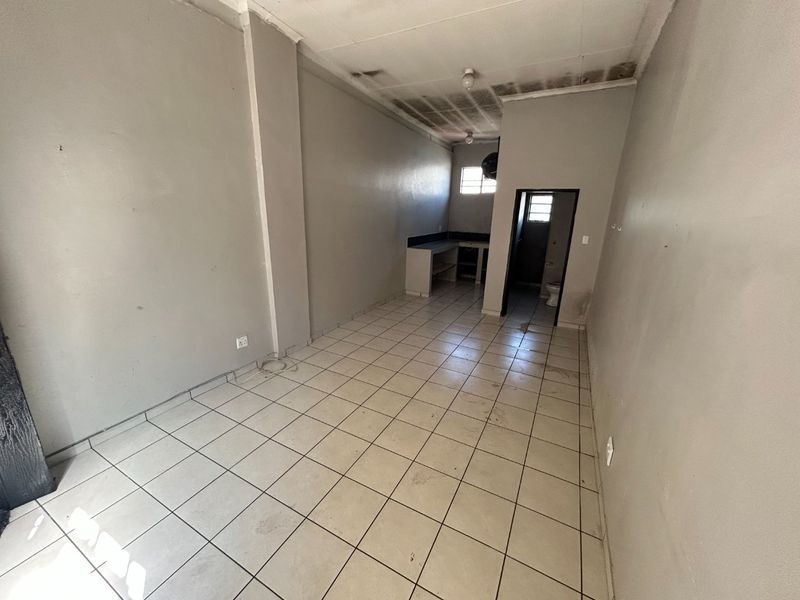 An empty open-plan commercial space featuring tiled flooring, a small kitchenette area, and an attached restroom, requiring cosmetic refurbishment and ceiling repairs.