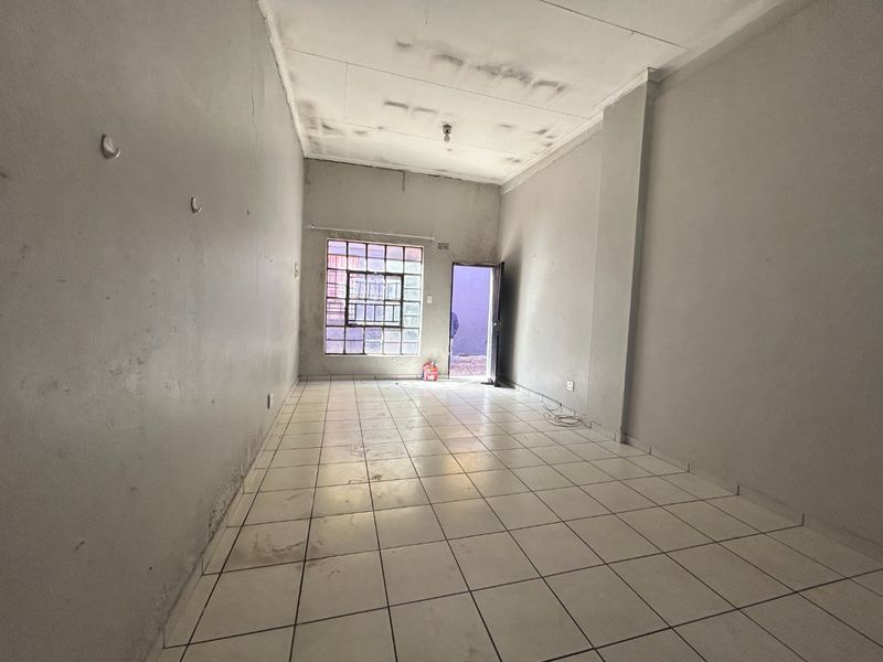 An empty, open-plan commercial space featuring tiled flooring, a steel-framed window with security bars, and a rear exit door.