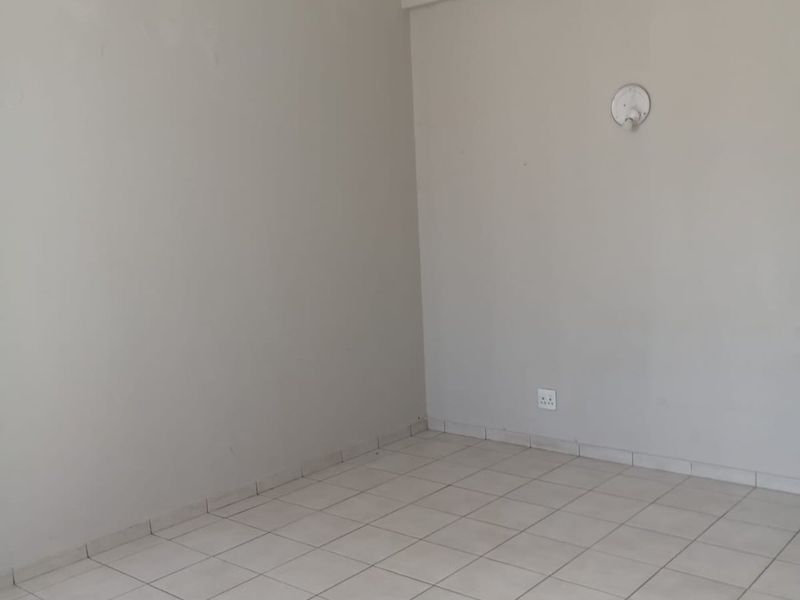 A vacant, neutral-toned office space featuring tiled flooring, a wall-mounted light fixture, and standard electrical outlets.