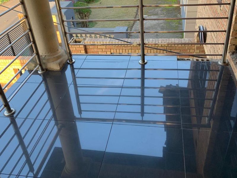 A clean, elevated balcony featuring modern dark floor tiles and a stainless steel balustrade, overlooking the front garden and street.