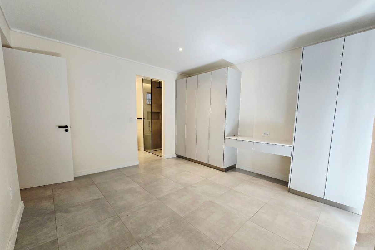 A bright, modern bedroom featuring neutral tiled flooring, extensive built-in cabinetry with an integrated desk, and direct access to an en-suite bathroom.