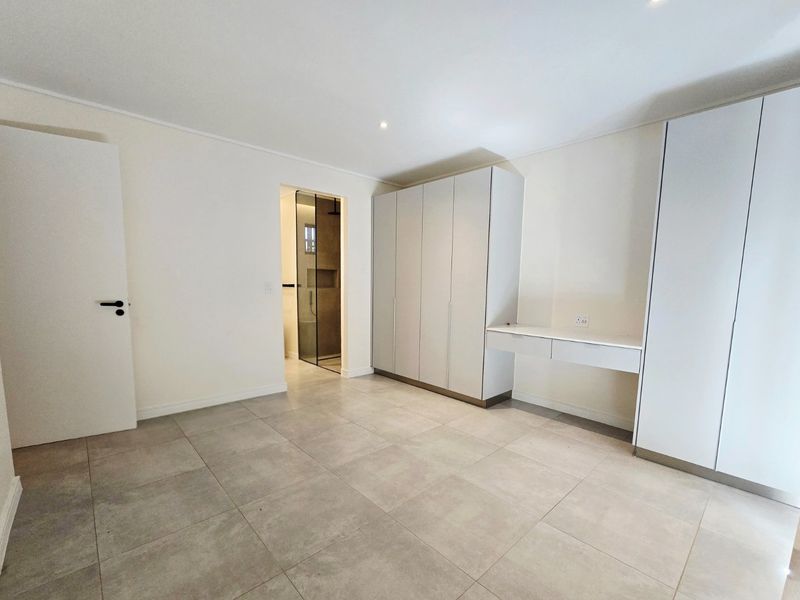 A bright, modern bedroom featuring neutral tiled flooring, extensive built-in cabinetry with an integrated desk, and direct access to an en-suite bathroom.