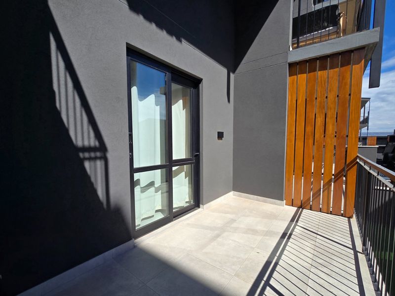A modern apartment balcony featuring tiled flooring, a timber privacy screen, and a sleek metal balustrade with access via a sliding glass door.