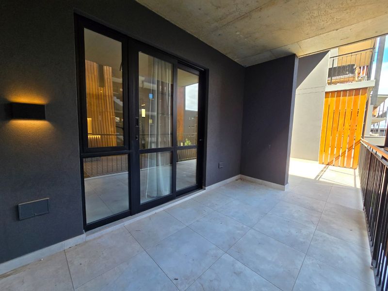 A modern, covered balcony featuring tiled flooring, exposed concrete ceiling, and sleek dark-framed sliding glass doors leading to the interior.