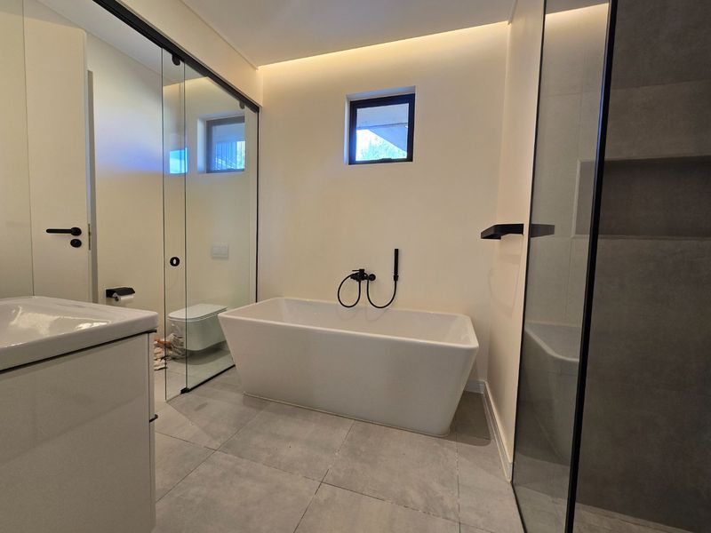Modern bathroom featuring a sleek freestanding bathtub, glass-enclosed shower, wall-mounted toilet, and contemporary vanity with high-quality finishes and ambient cove lighting.
