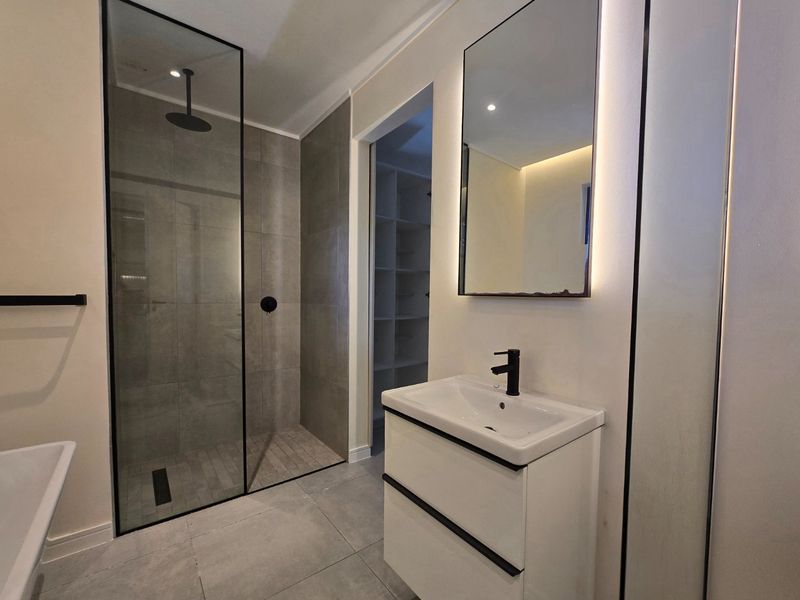 Modern bathroom featuring a walk-in shower with glass screen, contemporary vanity with basin, backlit mirror, and neutral floor-to-ceiling tiling.
