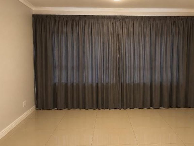 A spacious, empty living area featuring neutral tiled flooring, clean wall finishes, and a large window covered by floor-to-ceiling curtains.