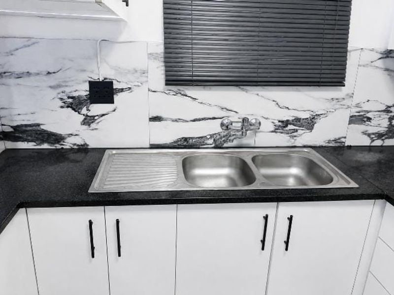 A clean, modern kitchen area featuring a stainless steel double sink, granite countertops, and marble-patterned splashback tiles.